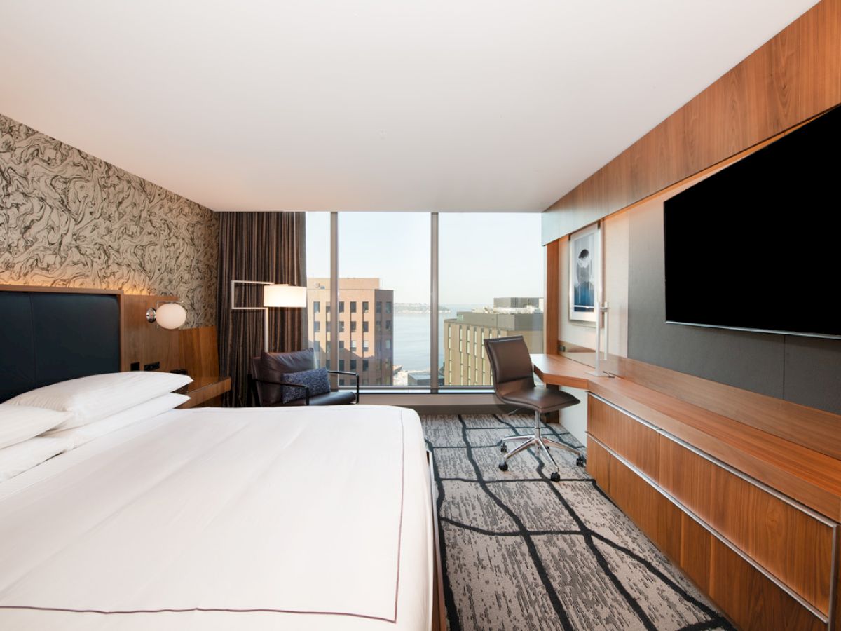 A modern hotel room with a large bed, desk, chair, TV, and a window view of buildings and water. The decor is sleek with patterned wallpaper.