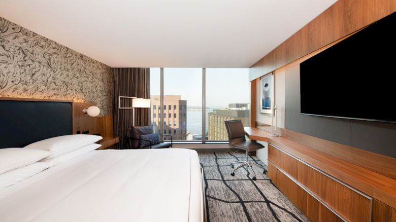 A modern hotel room with a large bed, desk, chair, TV, and a window view of buildings and water. The decor is sleek with patterned wallpaper.