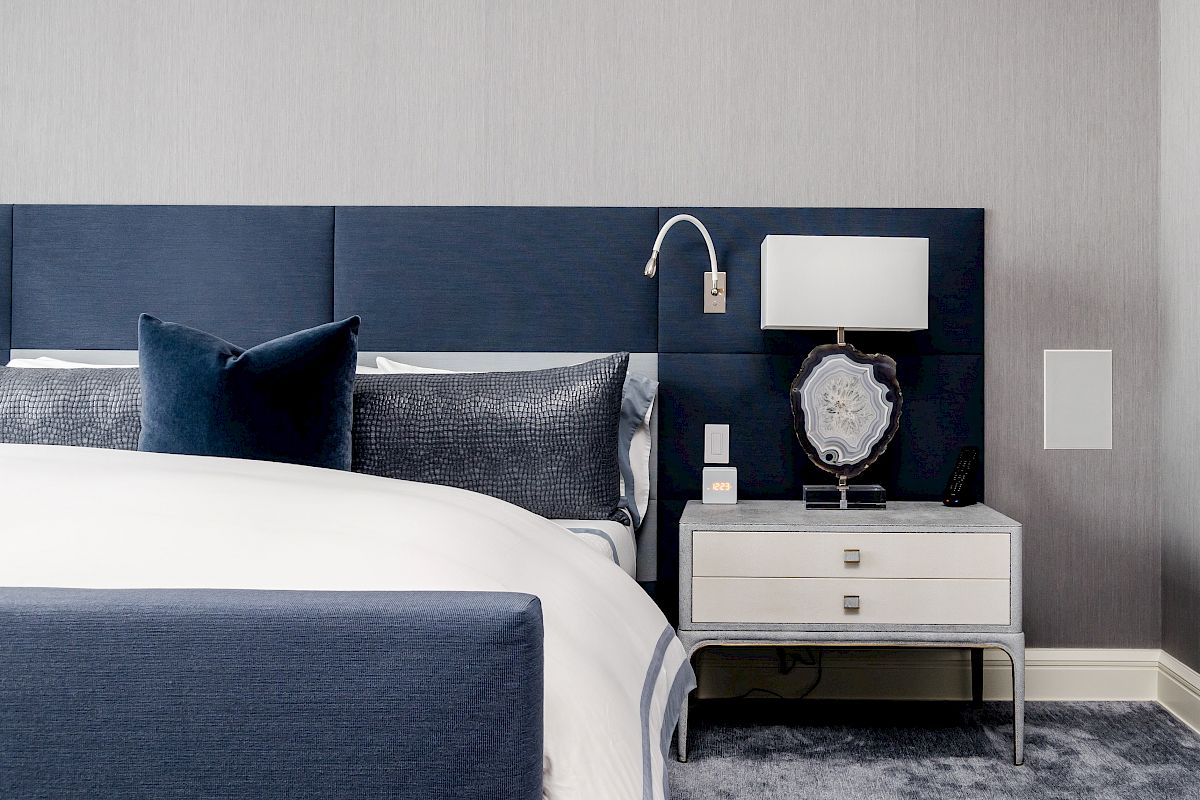 A modern bedroom features a navy blue upholstered bed, a white nightstand, and a lamp with an intricate base design.