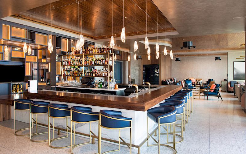 A modern bar with a wooden counter, blue chairs, hanging lights, and a well-stocked back shelf. Cozy seating in the background.